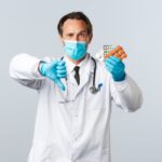 Covid-19, preventing virus, healthcare workers and vaccination concept. Displeased serious doctor in medical mask and gloves, show thumb-down and medication, bad quality pills, wrong prescription