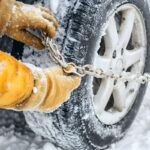 Hands attaching snow chains to car tires in snowy conditions, emphasizing winter safety, road preparedness, and essential cold weather driving equipment