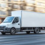 White Delivery Van Mockup Driving Through City Street, Template For Advertisement On Vehicle Side